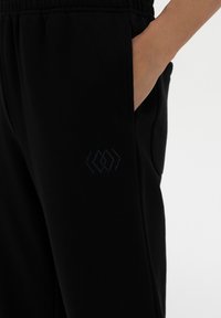 Black jogger pants with elastic waistband, side pockets, and tonal logo detail on the thigh. Made from soft, comfortable fabric.