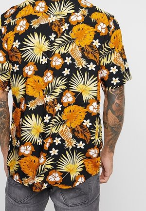 Chemise à manches courtes noire avec motif tropical de fleurs et d'ananas en orange, jaune et blanc portée par une personne tatouée.