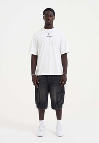 Overdimensioneret hvid bomulds t-shirt med sort logo, parret med sorte cargo shorts med store lommer, og hvide sneakers med teksturerede såler.