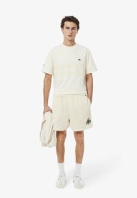 Tenue de sport couleur crème comprenant un t-shirt à manches courtes texturé et un short en maille, accentuée par un logo vert et des détails en bordure.