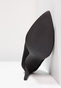 Svart mocka högklackad sko med spetsig tå och en slät svart sula med horisontella räfflor för bättre grepp. Positionerad mot en ljus bakgrund.