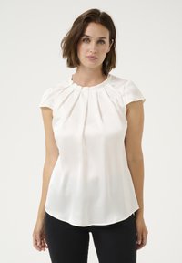 Cream short-sleeve blouse with pleated detailing at the neckline, smooth fabric, and a gently curved hem. Worn with black trousers.