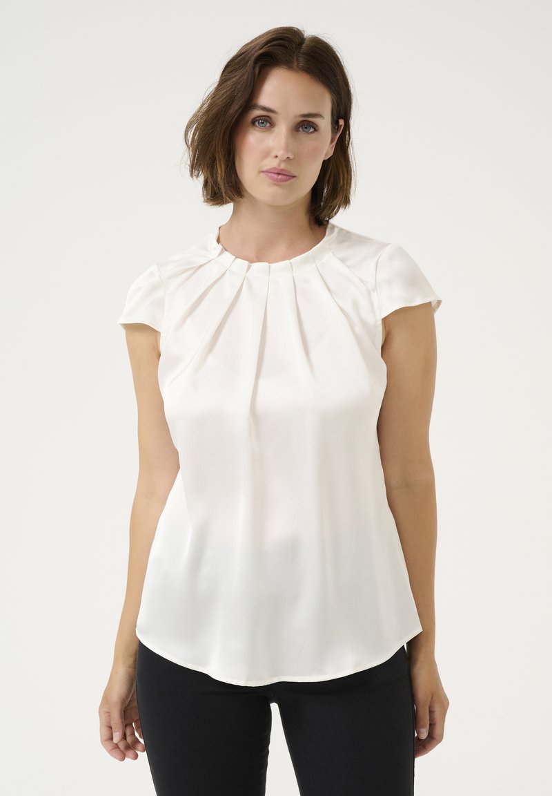 Cream short-sleeve blouse with pleated detailing at the neckline, smooth fabric, and a gently curved hem. Worn with black trousers.