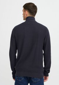 Sötét tengerészkék kötött pulóver magas gallérral, texturált mintával és bordás mandzsettákkal, amelyet kék farmernadrággal párosítanak.