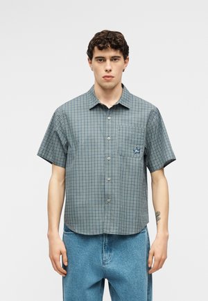 Mladi moški v kratkoremenski zeleni karirani srajci z gumbi in sprednjim žepom ter ohlapnih modrih kavbojkah stoji pred enostavnim ozadjem.