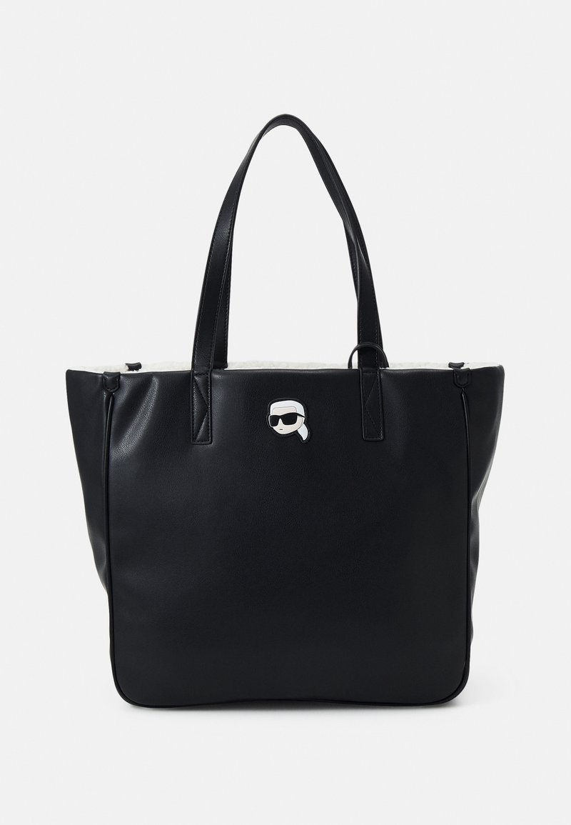 Sac cabas en cuir noir avec deux bandoulières et un petit patch blanc représentant une personne portant des lunettes de soleil et un foulard sur la tête à l'avant.