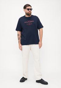 Granatowy bawełniany t-shirt z czerwonym graficznym logo, w zestawie z beżowymi spodniami i czarnymi butami. Model nosi okulary przeciwsłoneczne i ma brodę.