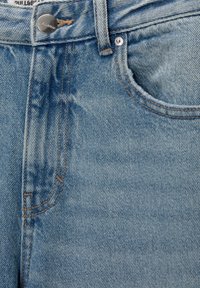 Gros plan sur un jean en denim bleu clair montrant le bouton, la passants de ceinture, les coutures et la poche avant avec une texture subtile du tissu.