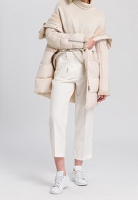 Personne portant un pull en tricot beige, un pantalon cropped de couleur claire, des baskets blanches et un manteau en doudoune beige drapé sur les épaules, sur fond uni.