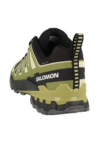 Scarpe da trail running verdi e nere con superficie testurizzata, suola robusta e dettagli riflettenti. Presentano una caviglia imbottita e un dettaglio con logo.