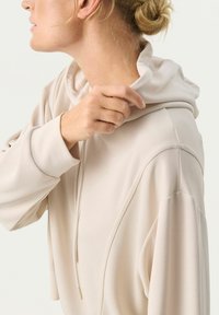 Pullover beige con cappuccio dal texture morbido e liscio. Presenta un collo alto e un cordino regolabile. Maniche ampie con cuciture sottili.