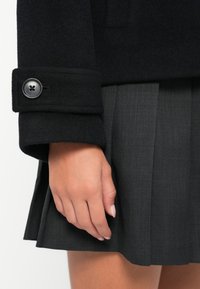 Kadar ruke pored tamnosive plisirane suknje i crne vune rukava s velikim crnim dugmetom.