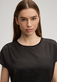 Top nero a maniche corte con tessuto liscio e trasparente, scollo rotondo e rifiniture nere a contrasto, che evidenziano un design semplice.