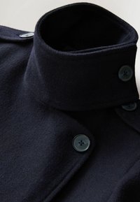 Manteau en laine bleu marine avec un col haut, comportant deux boutons foncés. Le tissu a une texture douce et un design soigné et ajusté.