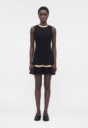 Γυναίκα με μαύρο αμάνικο φόρεμα με χρυσές λεπτομέρειες στο ντεκολτέ και το τελείωμα, στέκεται σε απλό φωτεινό φόντο.