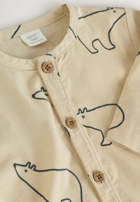 Beige babyromper i kordfloss med svarta djurmönster i kontur och bruna knappar med varumärket "NEXT" på framsidan.