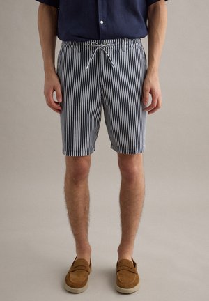 Homme portant une chemise bleu marine, un short rayé vertical bleu marine et blanc avec cordon de serrage, et des chaussures à enfiler couleur tan, debout sur un fond uni.