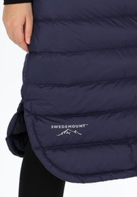 Laivastonsininen toppatakki, jossa on vaakasuoria tikkausosioita, pyöristetty helma ja valkoinen logo alaosassa. Materiaali näyttää sileältä ja eristetyltä.