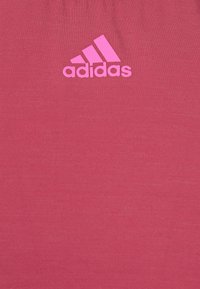 Vaaleanpunainen urheilutoppi, joka on valmistettu teksturoituneesta kankaasta ja jossa on näyttävä valkoinen Adidas logo etupuolella. Yksinkertainen, istuva malli.
