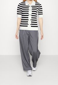 Maglione a righe bianche e nere a maniche corte con colletto e dettagli in bottone, abbinato a pantaloni larghi grigi e sneakers bianche.