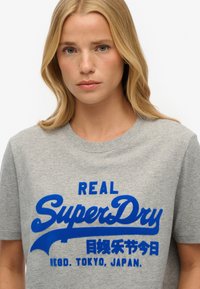 Szara bawełniana koszulka z odważnym niebieskim logo "SuperDry" oraz japońskim tekstem. Krótkie rękawy, okrągły dekolt i luźny krój.