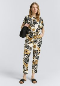 Zweiteiliges Outfit in Creme mit einem schwarz-braunen Blättermuster. Das Oberteil hat kurze Ärmel und die Hose ist tailliert mit einem elastischen Bund.