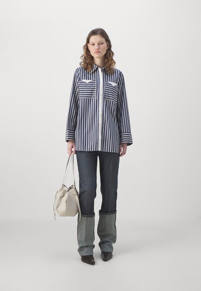 Camicia a righe bianca e blu navy con due tasche frontali, abbinata a jeans denim scuri. Il modello tiene in mano una borsa di colore beige chiaro.