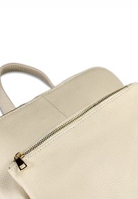 Crèmekleurige leren rugzak met een textuur, voorzien van een gouden rits en een handvat aan de bovenkant, zichtbaar tegen een witte achtergrond.