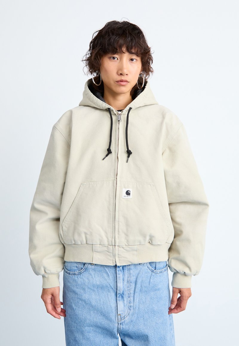 Person med kort krøllete hår iført en beige zip-up hettegenser og lyseblå jeans, stående mot en enkel lys bakgrunn.
