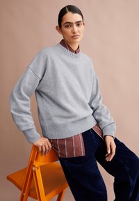 Frau mit nach hinten gekämmtem Haar trägt einen grauen Pullover über einem bordeaux-gestreiften Hemd und blauen Hosen und lehnt sich an einen leuchtend orangefarbenen Klappstuhl.