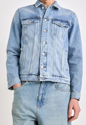 Person trägt eine hellblaue Jeansjacke mit Knopfverschluss und passende hellblaue Jeans, die Hände teilweise in den Taschen.