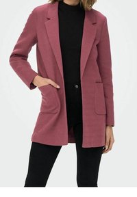 Veste rose à motif chevron avec deux poches frontales, fermeture à un bouton et col cranté, portée sur un haut noir.