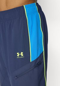 Shorts de sport navy en tissu léger présentent un panneau latéral bleu avec des accents néon et une poche zippée. Logo Under Armour affiché.