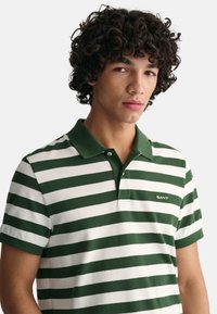Camisa polo a rayas verdes y blancas con un cuello verde sólido. Presenta un logo de GANT en la parte izquierda del pecho. La tela parece suave y texturizada.