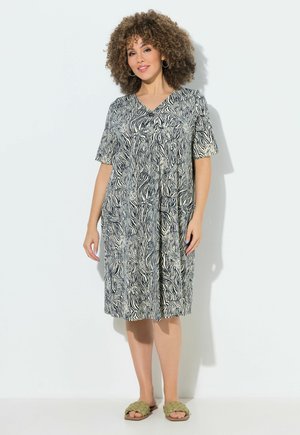 Frau mit lockigem Haar, die ein knielanges schwarz-weißes Kleid mit Zebramuster und grüne gewebte Slide-Sandalen trägt, steht vor einer weißen Wand.