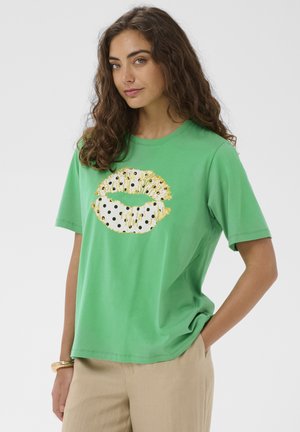 Femme aux longs cheveux bouclés portant un t-shirt vert à manches courtes avec un motif de lèvres à pois dorés et noirs, pantalon beige et bracelet, debout la main dans la poche.