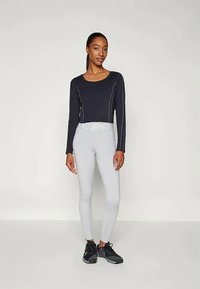 Crna sportska majica dugih rukava s bijelim šavovima, uparena sa svijetlosivim tajicama s teksturiranim pojasom i crnim sportskim cipelama.