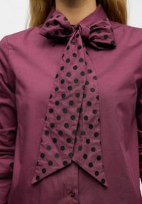 Sløjfeskjorte lavet af glat vinrødt stof med en stor sort polkaprikket sløjfe ved kraven og en lang, spids frontkant.