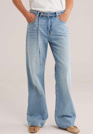 Personne portant un jean bleu clair taille haute à jambes larges avec une fine ceinture en denim, un haut blanc et des chaussures marron à enfiler avec un motif.