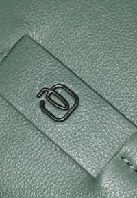 Cuero verde con textura y acabado suave, con un logo metálico negro en un diseño geométrico.