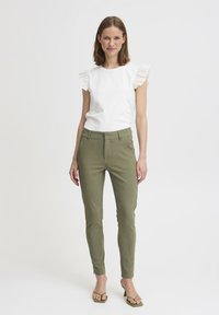 Haut blanc à manches courtes avec des dentelles aux épaules, associé à un pantalon slim-fit vert olive et des talons sandales verts. Design simple et épuré.