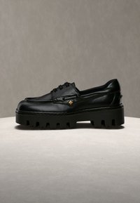 Mocassin à lacets en cuir noir avec semelle épaisse à crampons et petite décoration carrée dorée sur le côté, posé sur une surface claire avec un fond neutre.