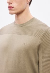 Beżowy sweter dziany z prążkowaną teksturą na górze, z subtelnym logo „BOSS” na piersi. Okrągły dekolt, gładka tkanina.