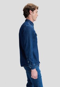 Vyras šonu įsikūnijęs, dėvi tamsiai mėlyną džinsinę marškinius ir džinsus, stovi priešais paprastą šviesiai pilką foną.