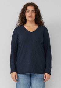 Frau mit lockigem Haar, die ein navyblaues langärmliges V-Ausschnitt-Shirt und hellblaue Jeans trägt, vor einem schlichten grauen Hintergrund steht.