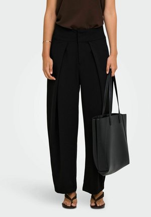 Femme portant un pantalon noir large et un haut marron, tenant un sac cabas en cuir noir, debout en sandales sur fond blanc.