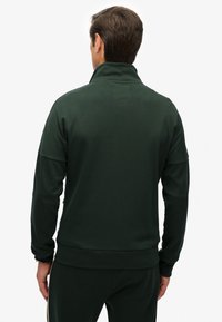 Chaqueta deportiva verde oscuro con cuello alto, confeccionada en tela lisa. Presenta un ajuste relajado y puños acanalados, combinada con pantalones a juego.