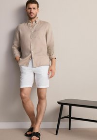 Beige Leinenhemd mit Knöpfen, weiße Shorts und schwarze Sandalen. Das Hemd hat eine Brusttasche, aufgerollte Ärmel und einen lässigen Schnitt.