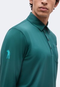 Homme portant un polo à manches longues bleu sarcelle avec un petit logo de silhouette bleu clair sur la manche gauche.