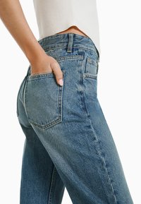 Jeans de mezclilla azul con un corte relajado, cintura de altura media y bolsillos traseros cosidos. Textura visible y áreas descoloridas, con un acabado suave y liso.